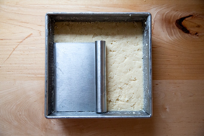 A square pan of unbaked shortbread with a bench scraper on top.